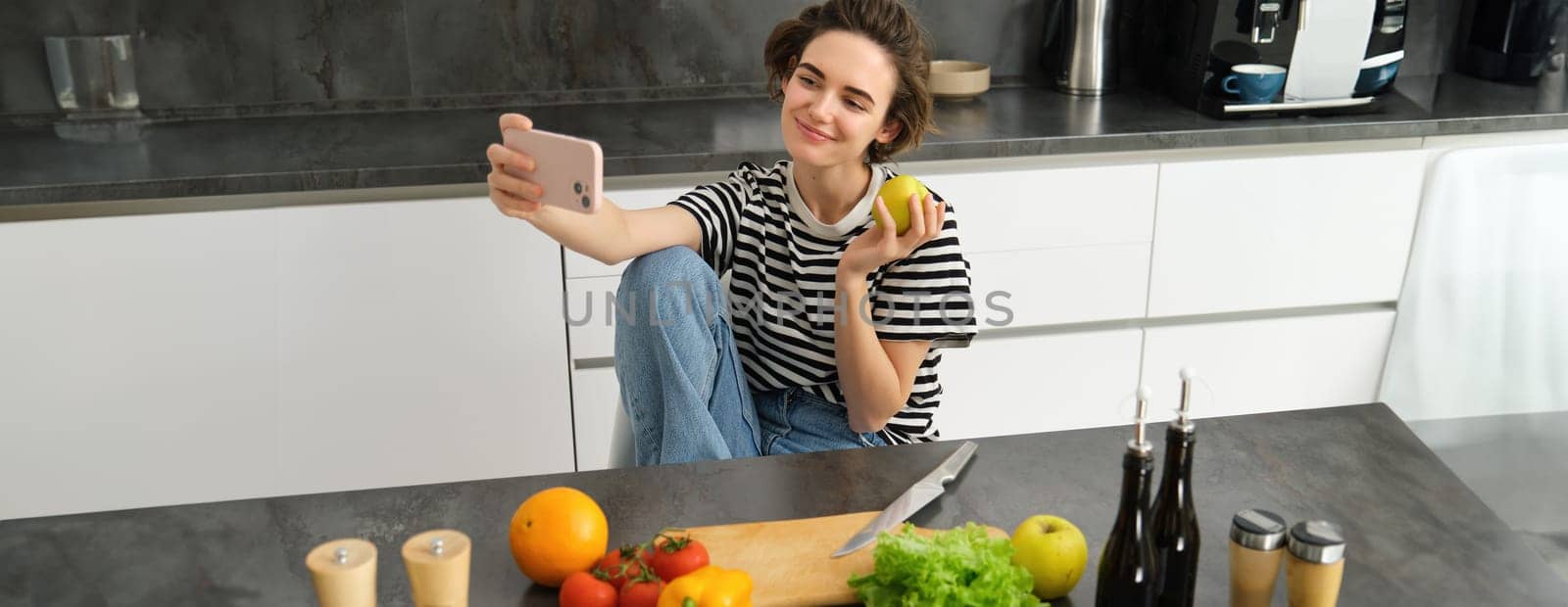 Portrait of smiling brunette woman in the kitchen, posing with an apple, taking selfie on smartphone, cooking and making pictures for internet blog.