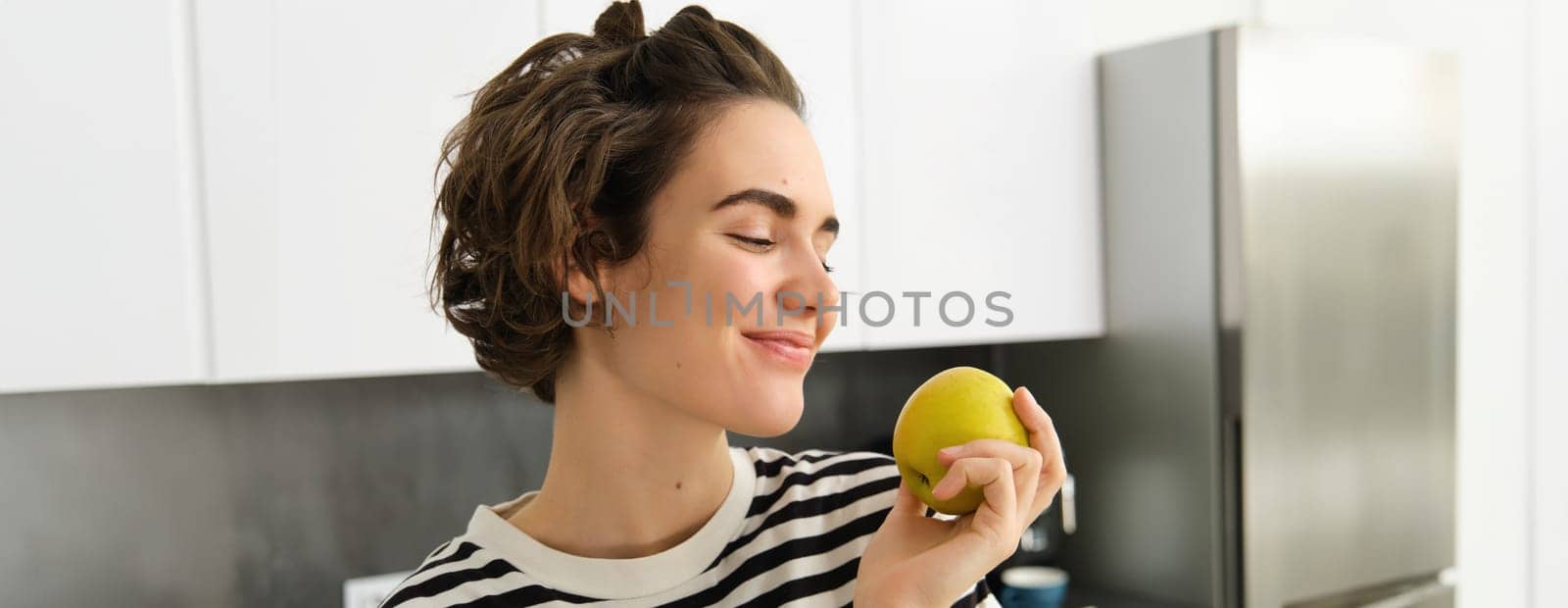 Close up portrait of beautiful, smiling brunette woman, holding an apple with pleased face and closed eyes, standing in the kitchen.