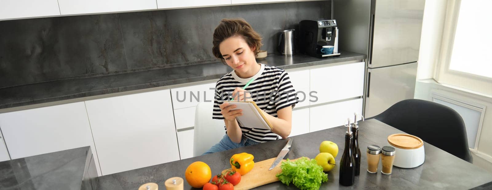 Portrait of woman writing down her recipe, making notes in notebook during cooking, sitting in the kitchen with chopping board and vegetables, preparing a meal by Benzoix