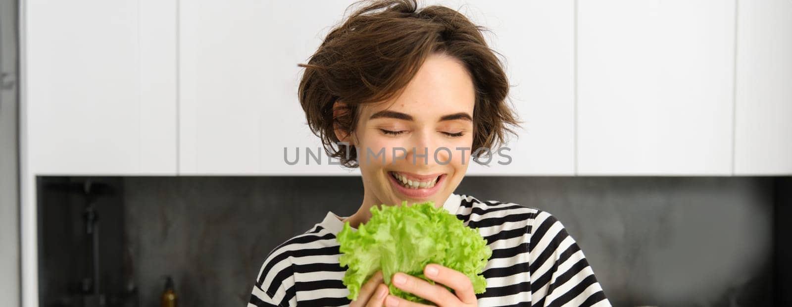 Portrait of happy and healthy young woman, following her diet, posing with lettuce leaf and smiling, cooking in the kitchen, vegetarian loves her vegetables by Benzoix