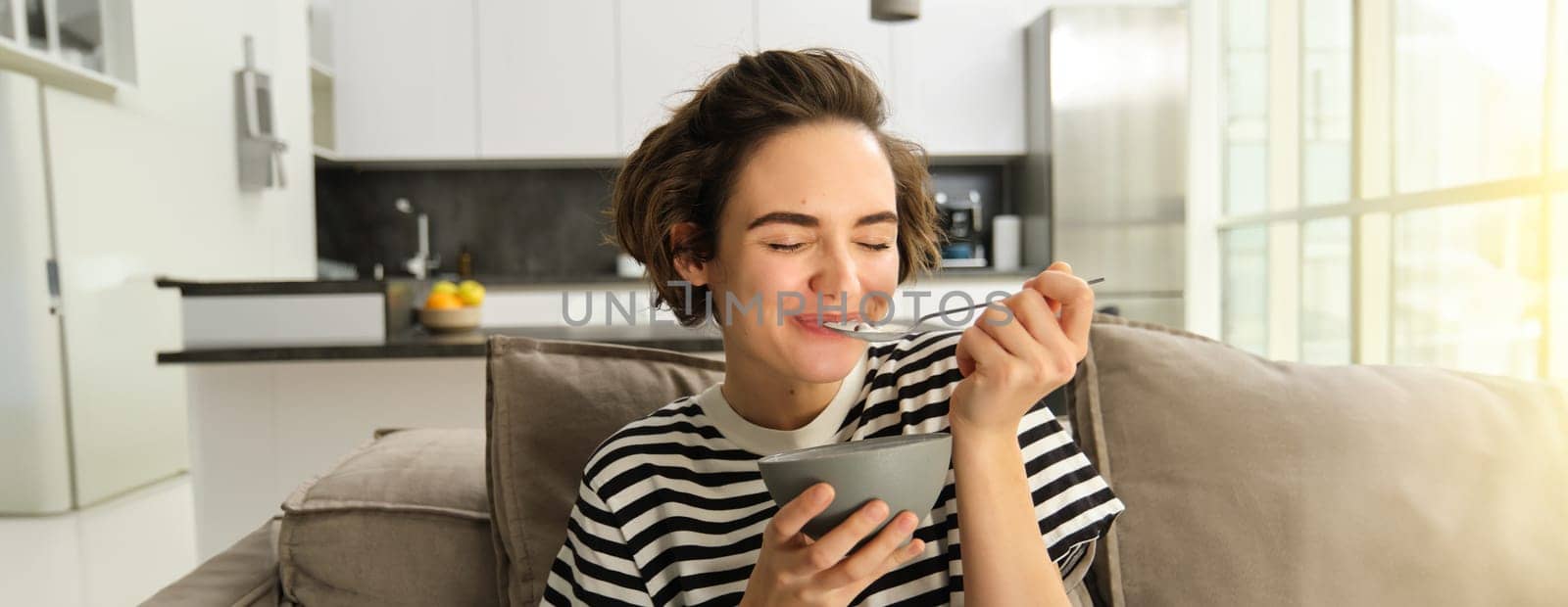 Portrait of happy brunette woman sitting on sofa and enjoying her breakfast, eating cereals, healthy granola in bowl, spending time in living room.