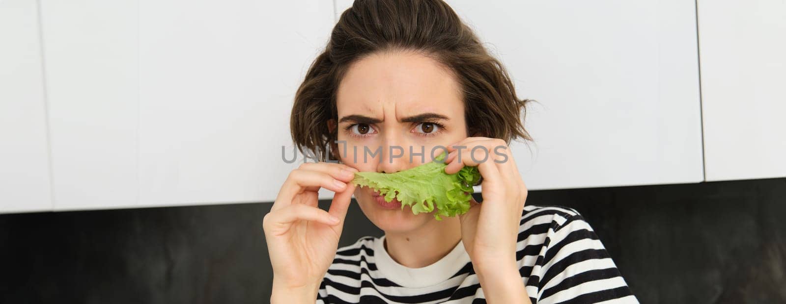 Frowning silly woman, holding lettuce leaf near face and frowning at camera, making pouting face, dislike eating vegetables.