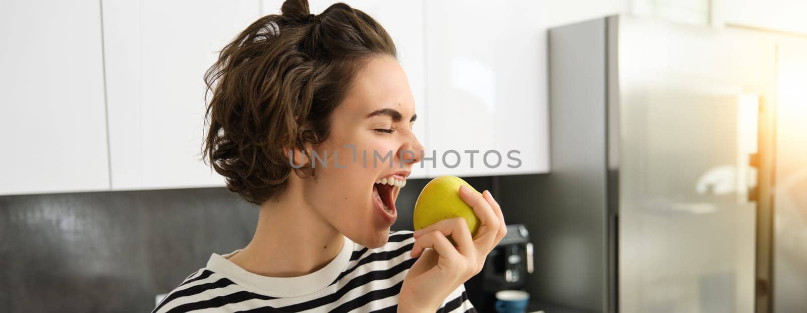 Close up portrait of smiling woman in the kitchen, holding an apple, eating fruit, having a healthy snack for lunch at home.