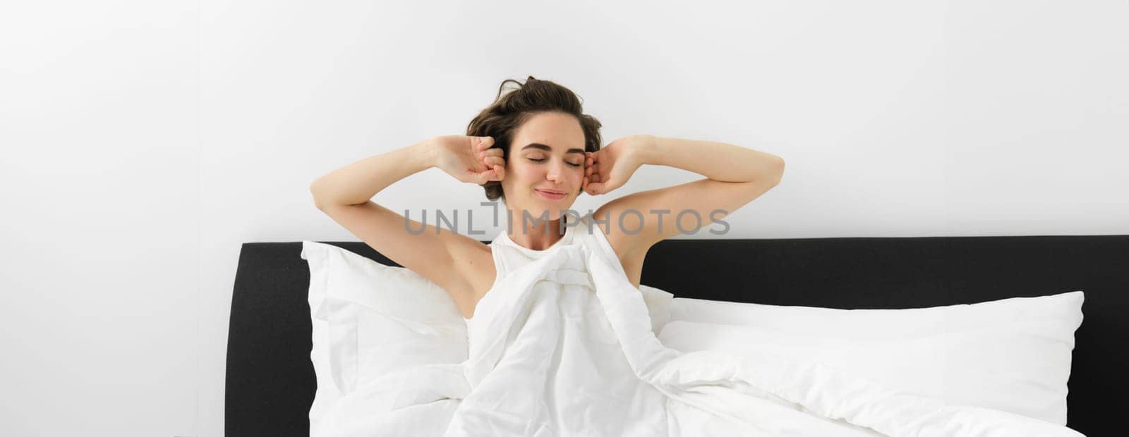 Beautiful young woman waking up in her bedroom, stretching hands with sleepy smiling face, sitting in bed under white linen duvet, had a good night sleep.