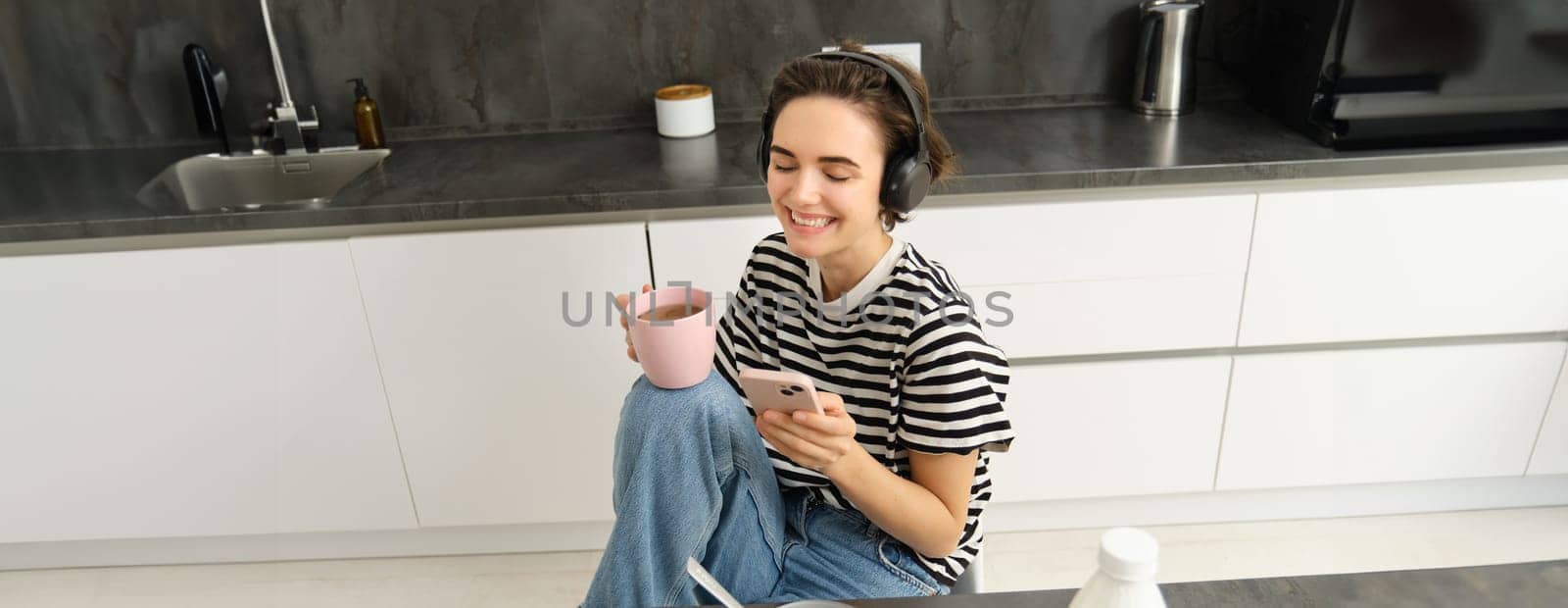 Portrait of young woman drinking tea and listening to music in headphones, scrolling social media while having lunch in kitchen, smiling happily.