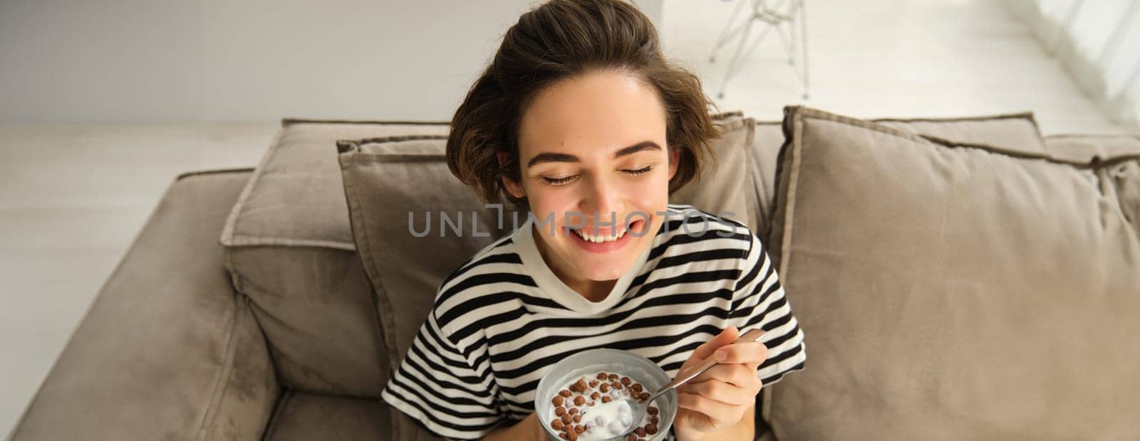 Upper angle shot of happy, cute young woman on sofa, eating bowl of cereals with milk and smiling, enjoying her breakfast.