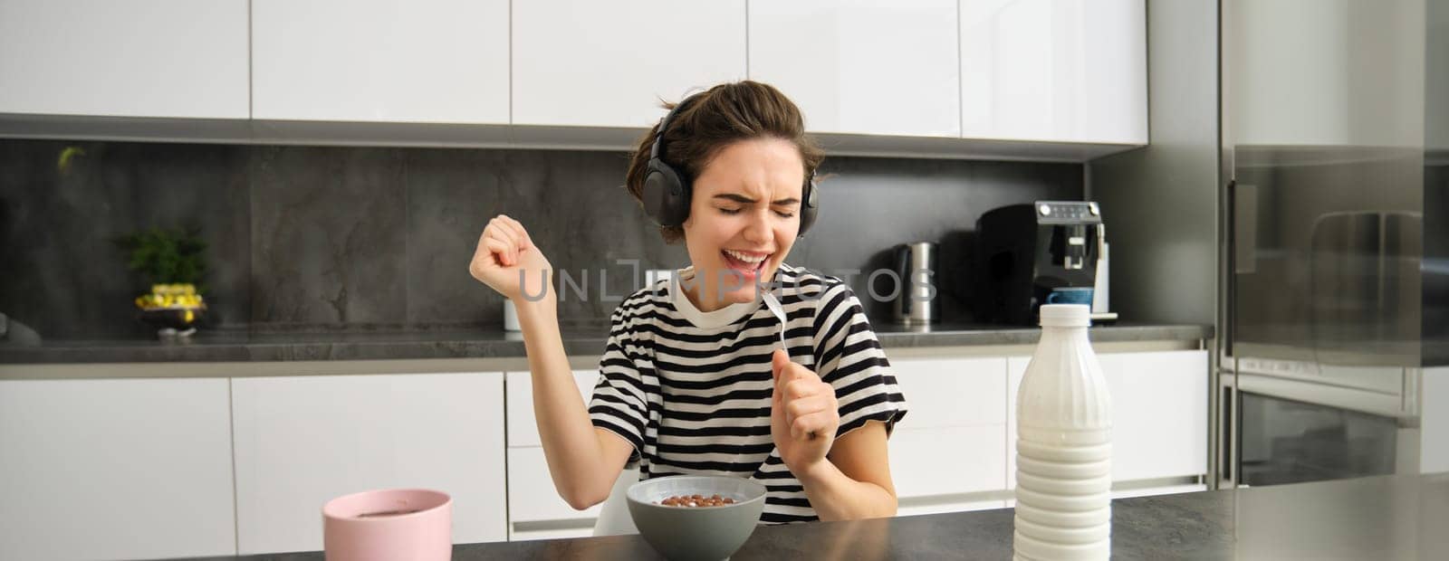 Happy and emotional young woman, singing while eating breakfast, having her cereals with milk, listening to music in wireless headphones, sitting in the kitchen.