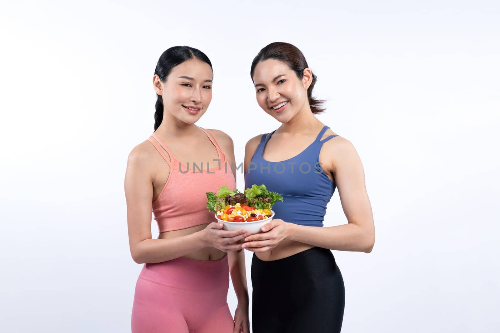 Two young sporty Asian women in sportswear holding salad bowl fill with fruit and vegetable. Natural youthful and fit body lifestyle people with balance nutrition on isolated background. Vigorous