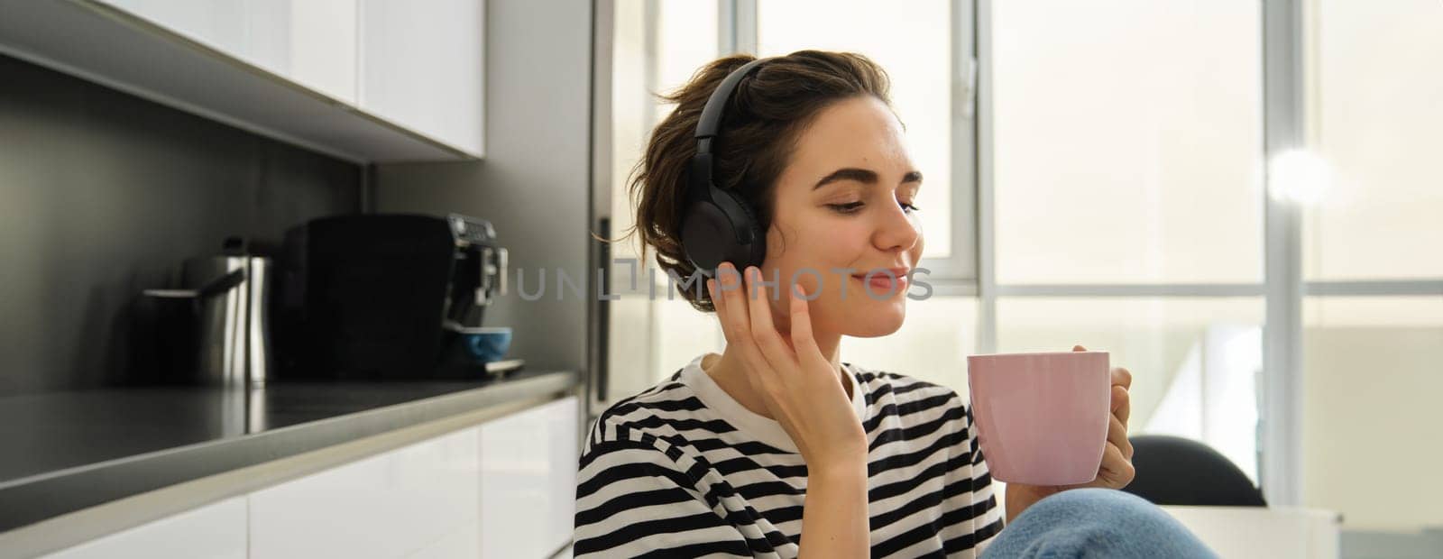 Close up portrait of smiling brunette woman, student in headphones, listens music, drinks cup of tea in morning, sits in kitchen and enjoys her daily routine.