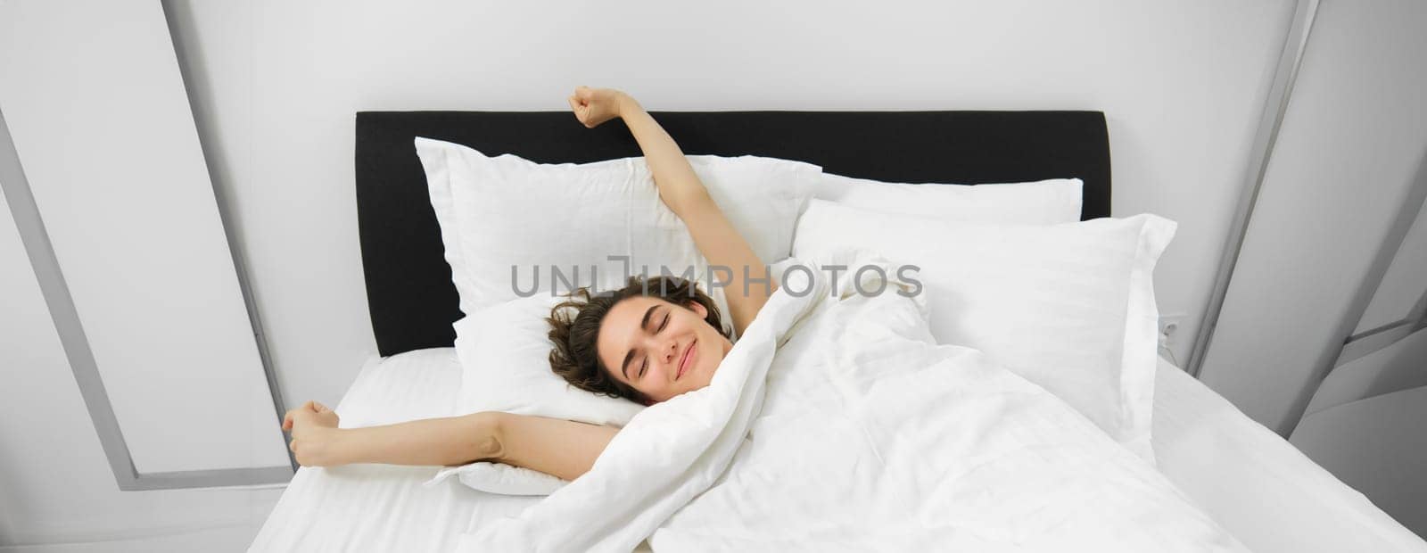 Portrait of happy, pleased young woman, stretching her arms with satisfied smile, lying in bed of soft pillow, waking up alone in bed, had good night sleep.