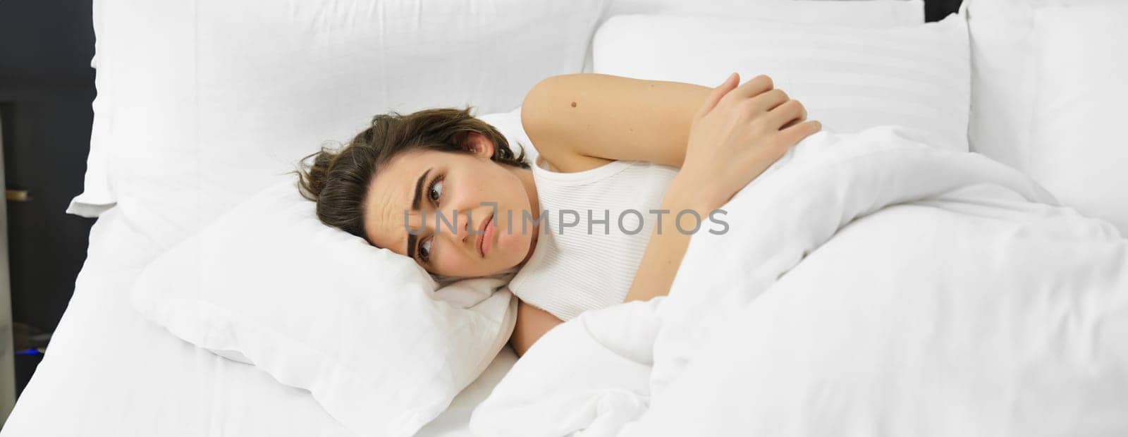 Portrait of woman feeling unwell, lying in bed with menstrual pain, stomach ache, frowning and looking sad. Copy space