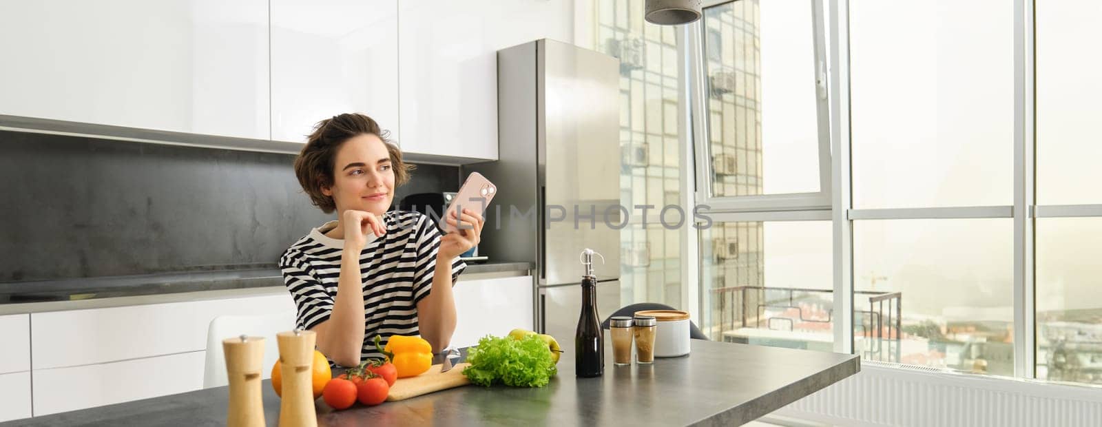 Portrait of young woman standing in the kitchen with vegetables and chopping board, holding smartphone, searching healthy diet recipes on mobile phone browser, cooking at home.