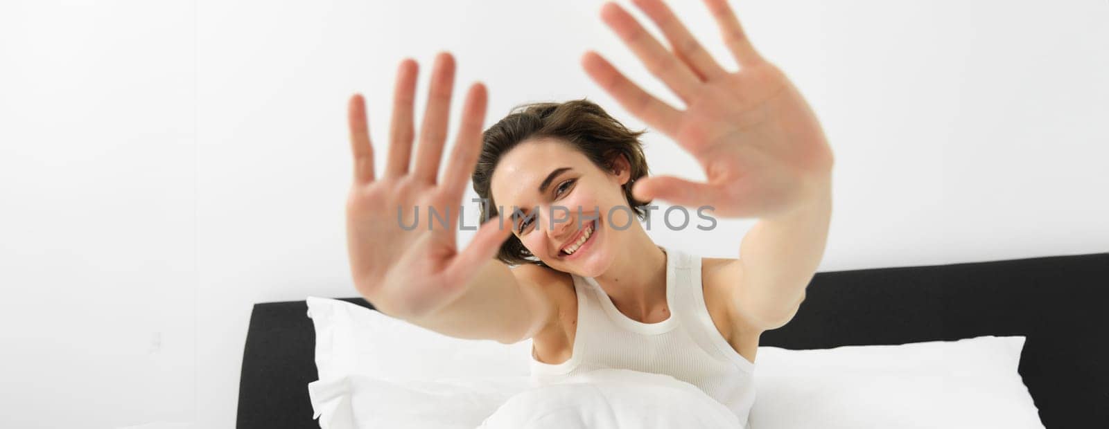 Portrait of sleepy, happy young woman, wakes up in her bed, stretching out hands, hiding face from camera, smiling and laughing.