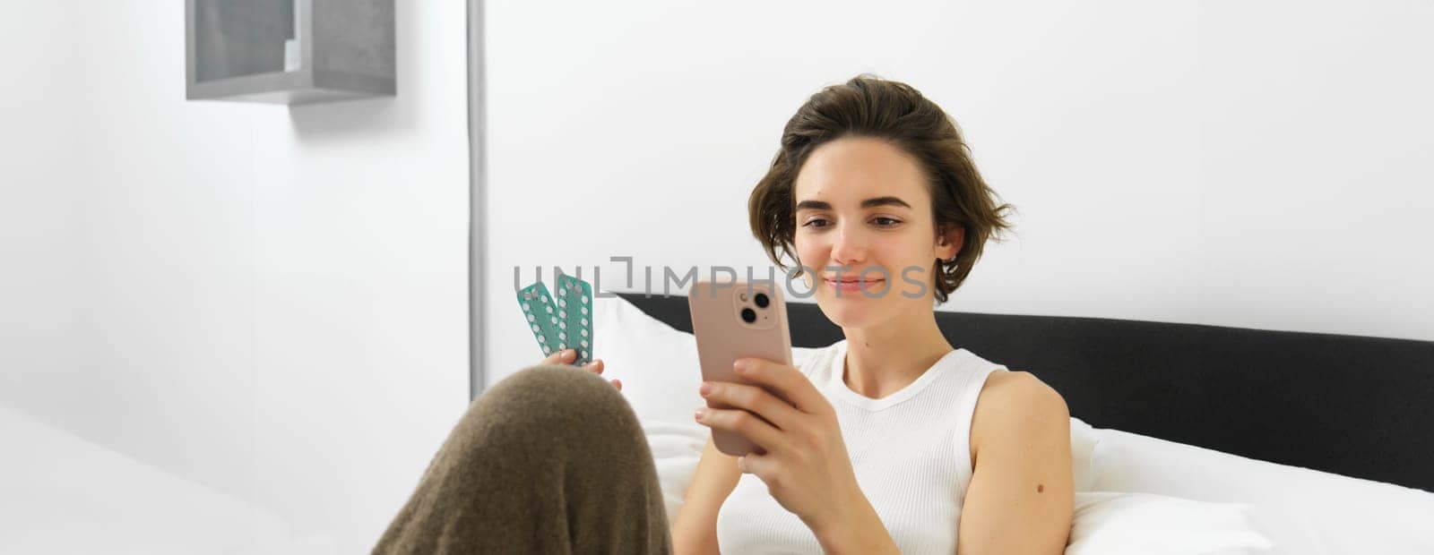 Woman searching for dosage instruction, holding tablets, her medication in hand, looking at mobile phone, lying in bed and smiling.