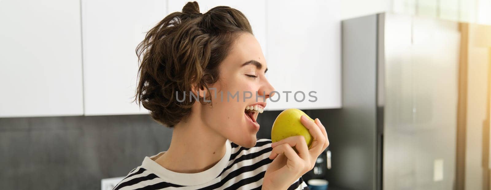 Close up portrait of young brunette woman biting an apple with pleasure, has pleased smile on her face, standing in the kitchen, having healthy snack for lunch, eating fruits.