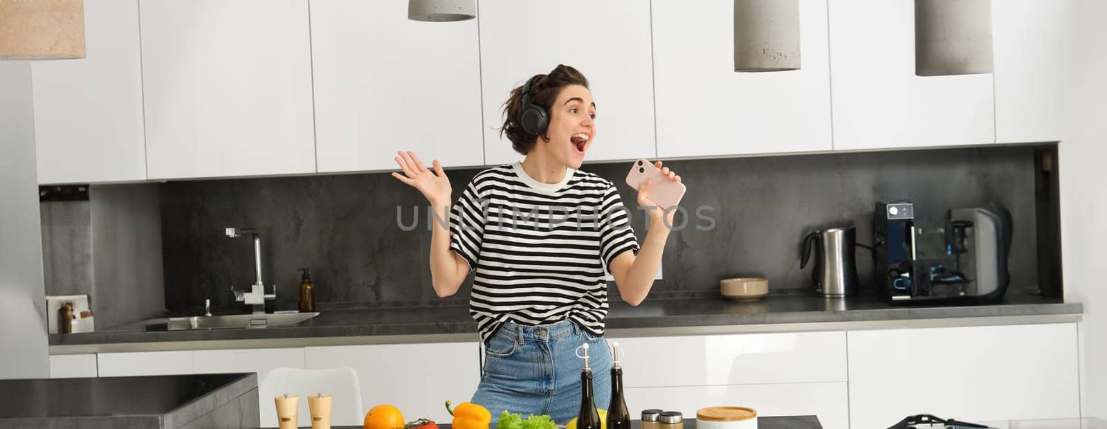 Portrait of carefree woman singing in smartphone mic, listening music in headphones while cooking breakfast, making a meal, chopping vegetables and dancing.