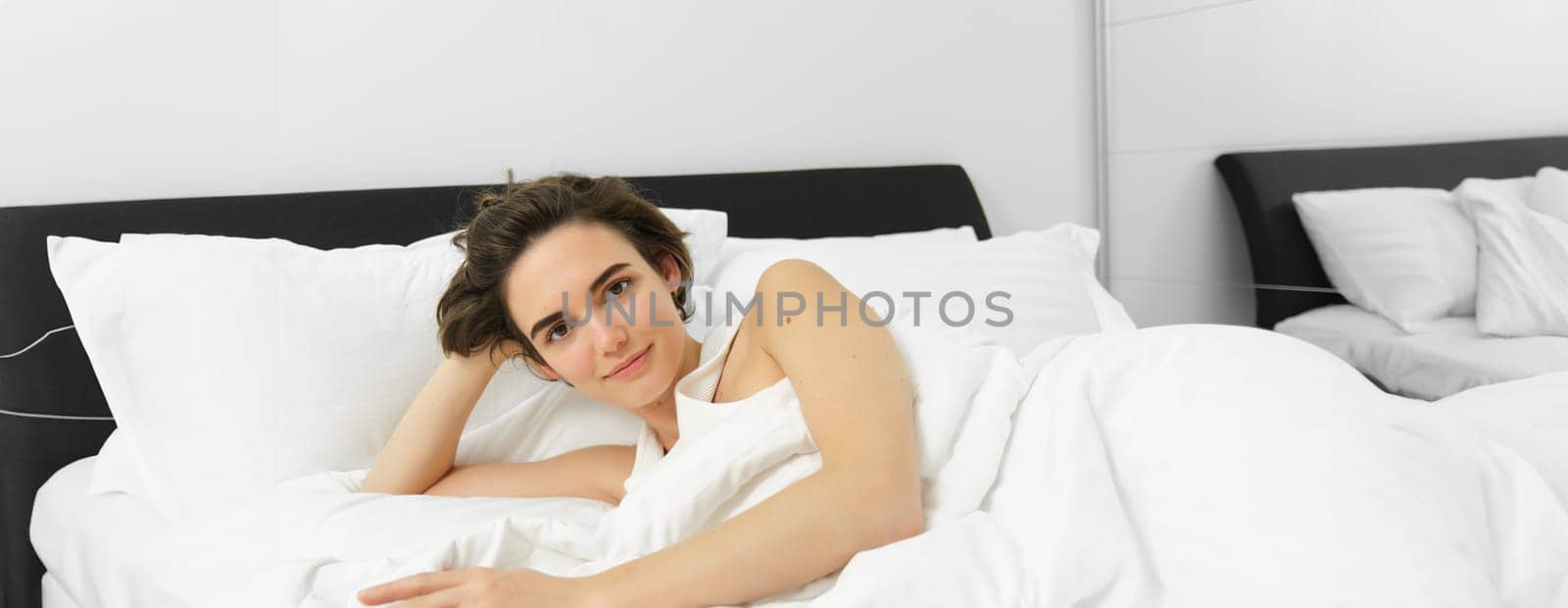 Portrait of beautiful woman in bed, waking up in morning, relaxing under white blanket with comfortable and soft pillows, looking happy and pleased.