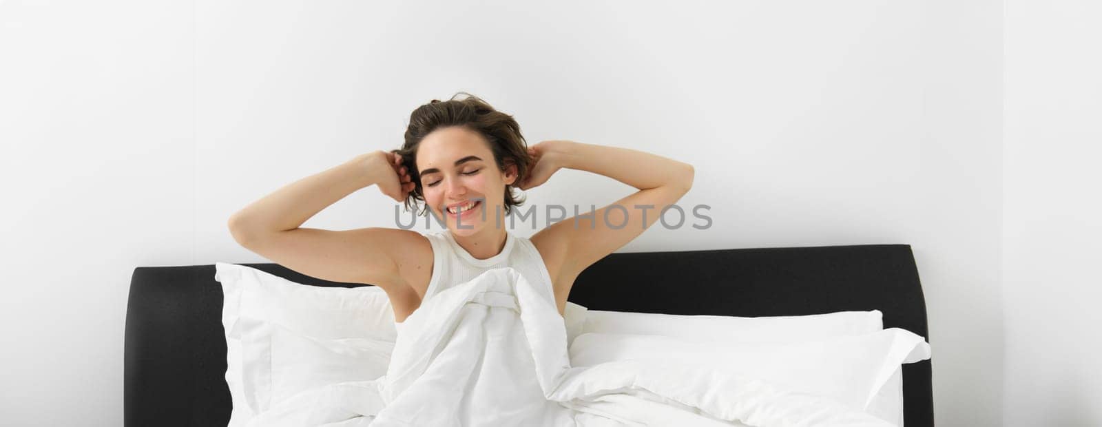 Portrait of woman feeling satisfied after good sleep, stretching hands after waking up, sitting on bed under white linen blanket, relaxing at home.