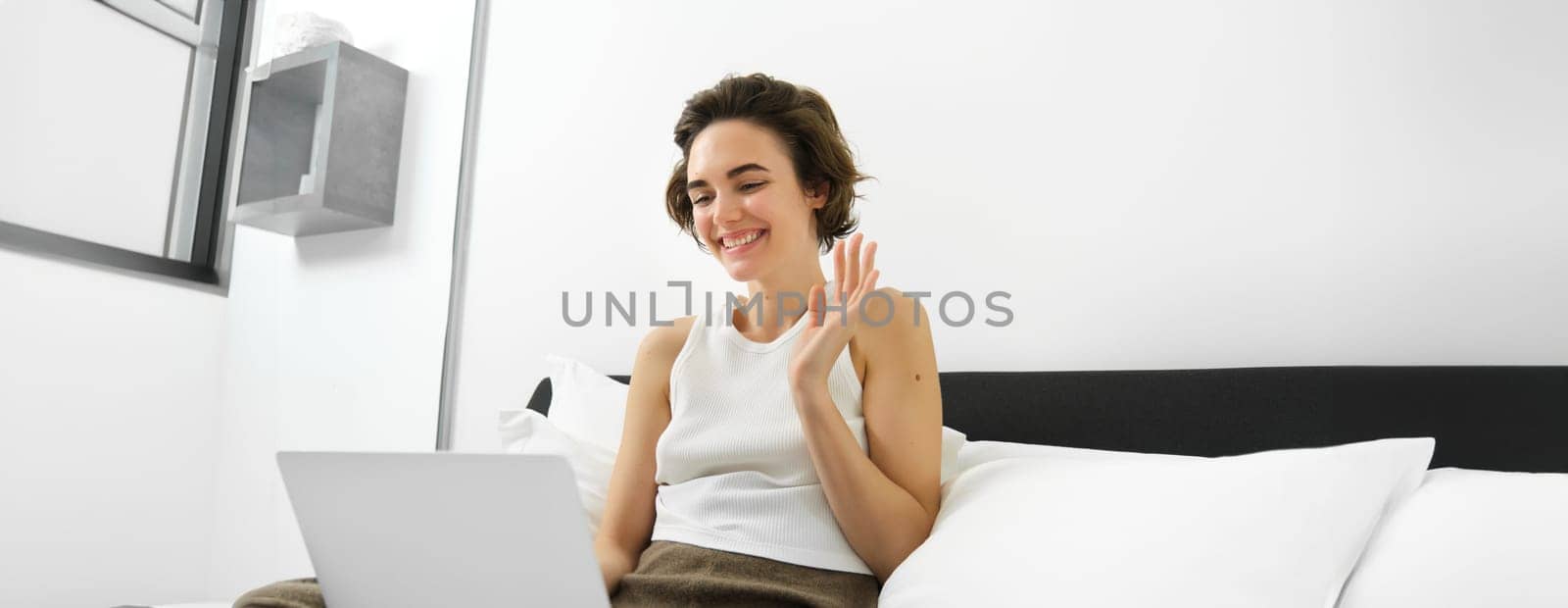 Smiling young woman, student connects to online lesson, video chats on laptop, waves hand at computer camera, says hello, greets someone, sits on bed in bedroom at home.