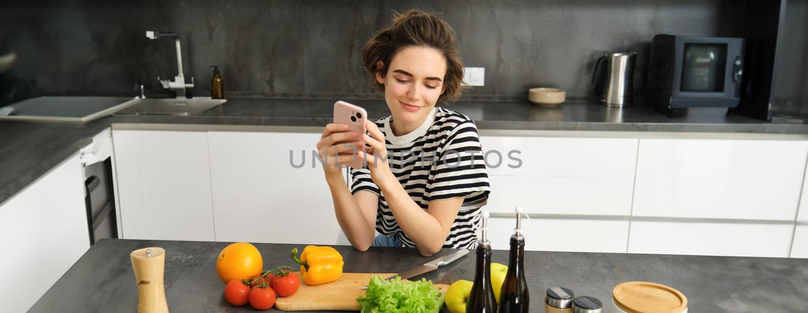 Portrait of young woman with smartphone, sitting in the kitchen and cooking salad, searching for healthy recipe online, has vegetables and chopping board on the table.