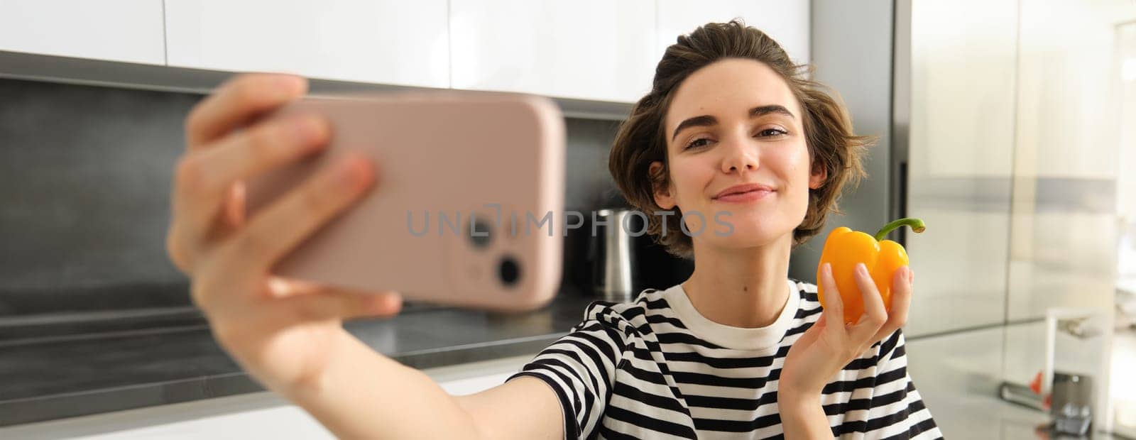 Smiling, beautiful young woman taking selfie with fresh yellow pepper, food blogger taking selfie with vegetables in the kitchen, vegan making a meal and recording video for social media.