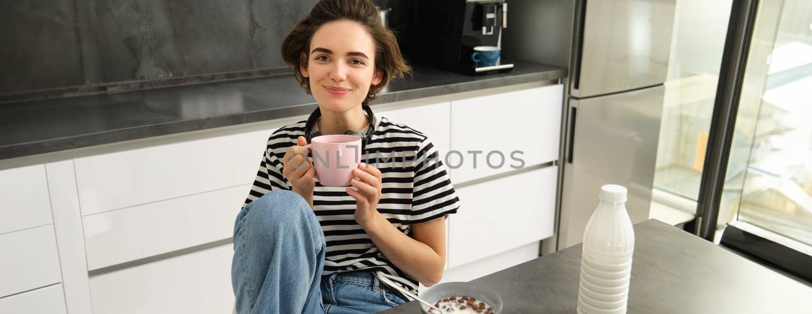 Cute brunette woman, student eating breakfast in morning, drinking hot tea, smiling and looking pleased.