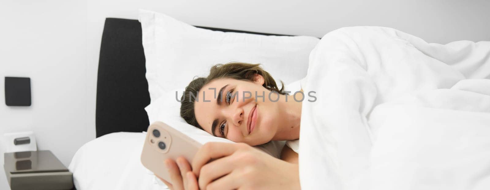 Close up portrait of lovely young woman in bed, lying and going to sleep, checking her phone messages, using smartphone and smiling.