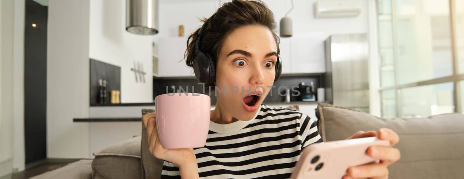 Portrait of woman binge watching her favourite tv show on smartphone, drinking tea and wearing headphones, gasping from shock, looking amazed at mobile screen.