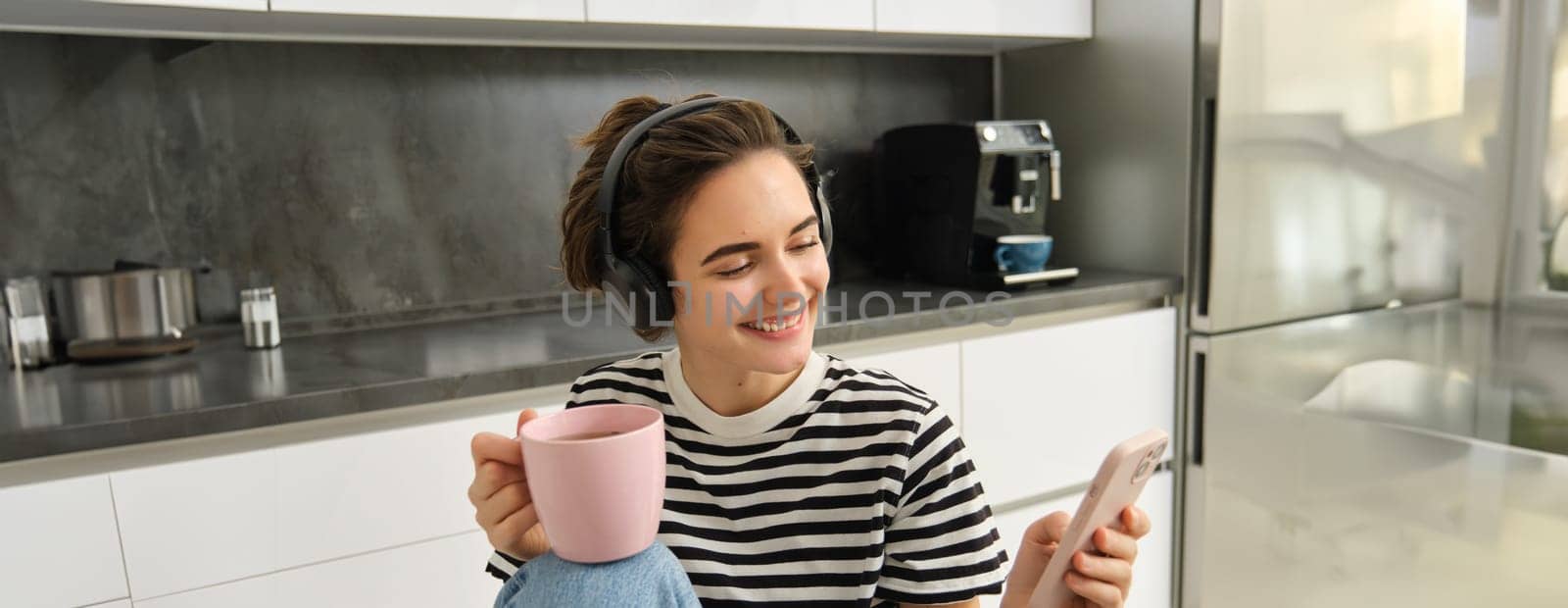 Portrait of young woman drinking tea and listening to music in headphones, scrolling social media while having lunch in kitchen, smiling happily.