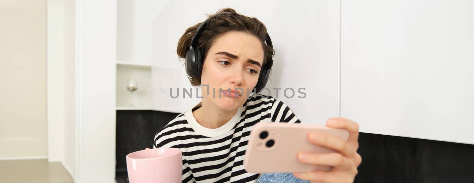 Portrait of woman with sad face, watching something on mobile phone, social media app, drinking tea, wearing wireless headphones.