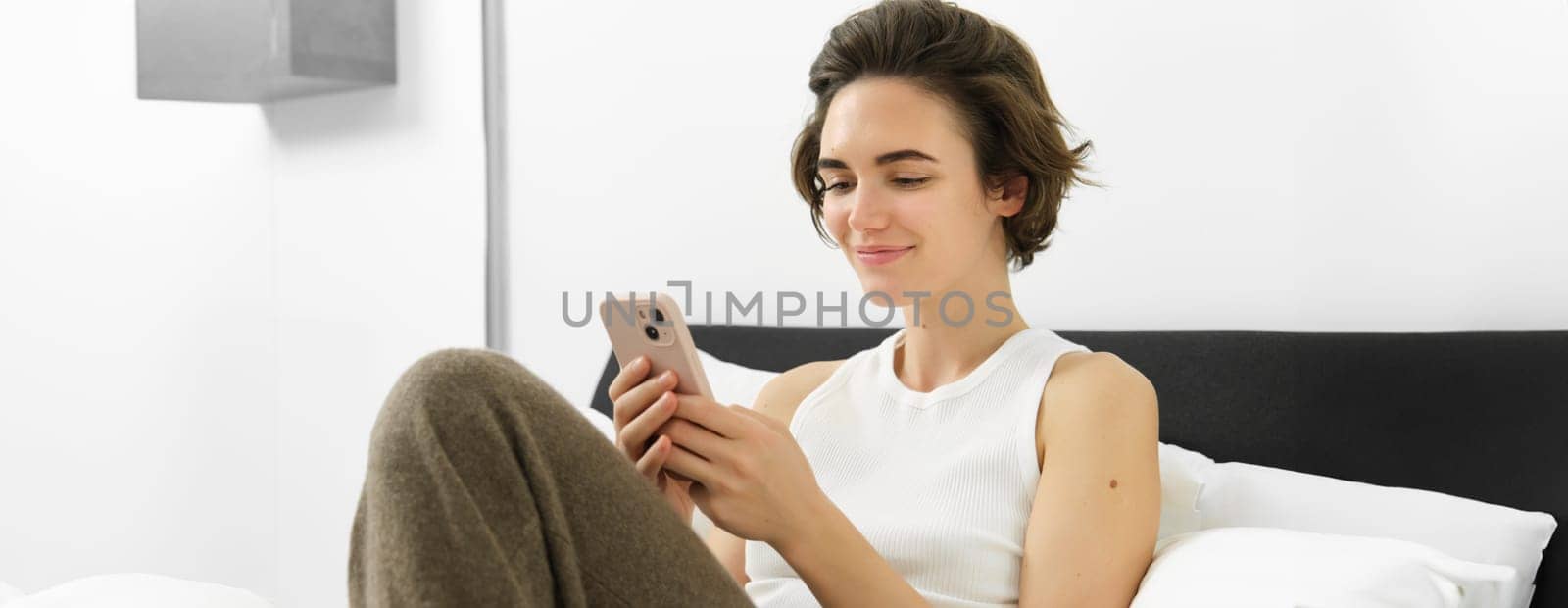 Cute young woman with mobile phone in hands, lying in bed in tank top, looking at smartphone with pleased smile.