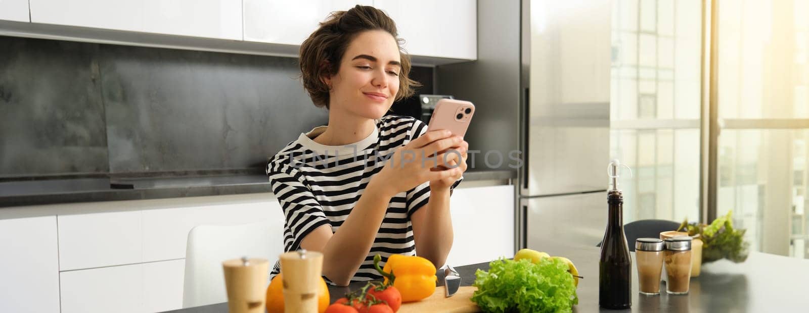 Portrait of young woman food blogger, posting on social media recipe, vegan salad, standing in the kitchen with smartphone, chopping vegetables.