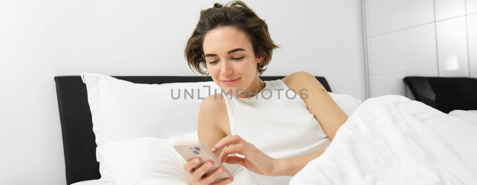 Image of female model in bed, looking at smartphone, sending a message, turns on alarm clock on mobile phone, goes to sleep in her bedroom.