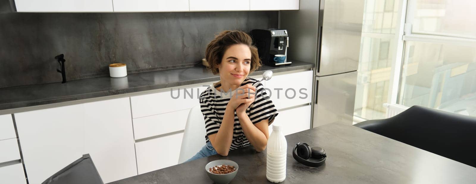 Portrait of smiling young candid woman, student eating morning cereals, having her breakfast, listening music in wireless headphones, sitting in the kitchen.