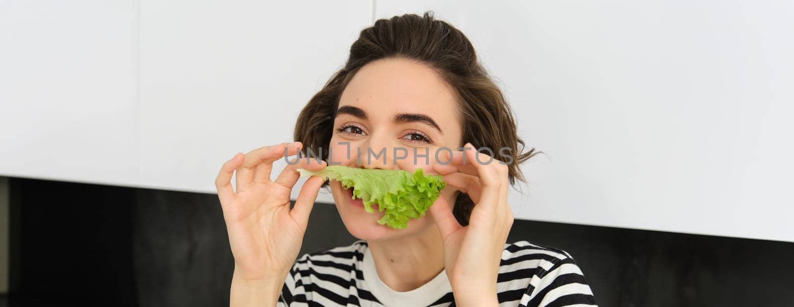 Close up portrait of young woman, vegetarian girl, likes eating vegetables, posing with lettuce leaf and smiling, posing in the kitchen. Concept of healthy food and diet.