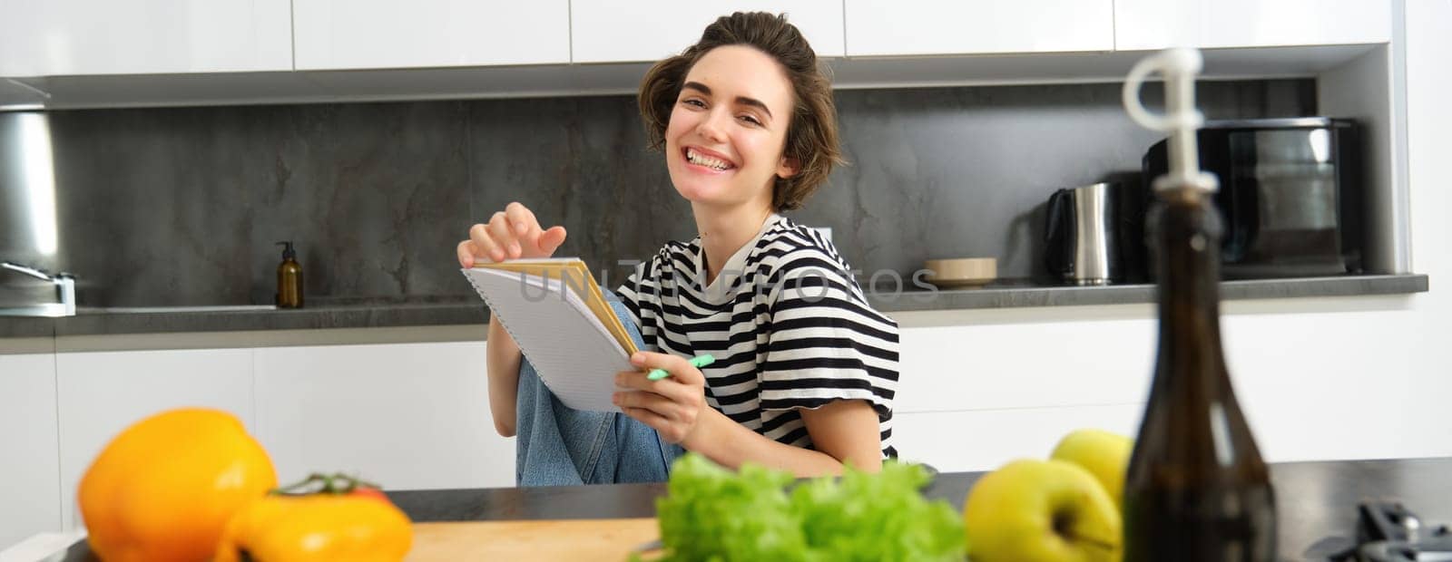 Portrait of young woman cooking, writing notes, grocery list in notebook, creating list of meals to cook through meal, sitting in kitchen near vegetables and chopping board.