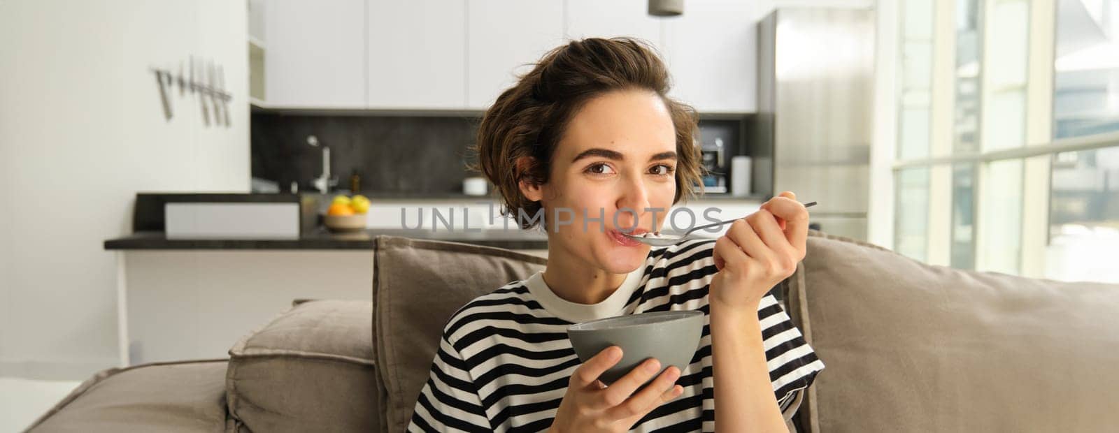 Portrait of young woman eating granola, bowl of cereals with milk, sitting on sofa and having her breakfast.