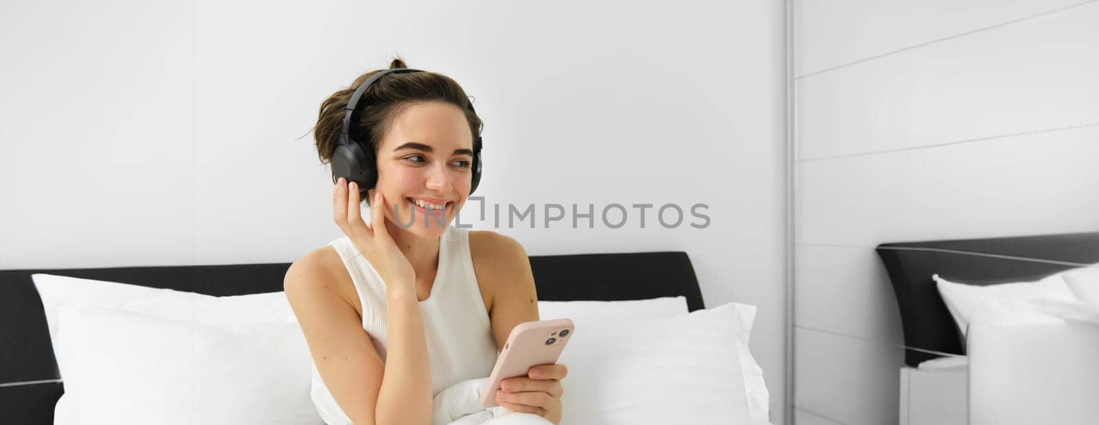 Beautiful girl in bed, listens to music in wireless headphones, using smartphone and smiling. Copy space