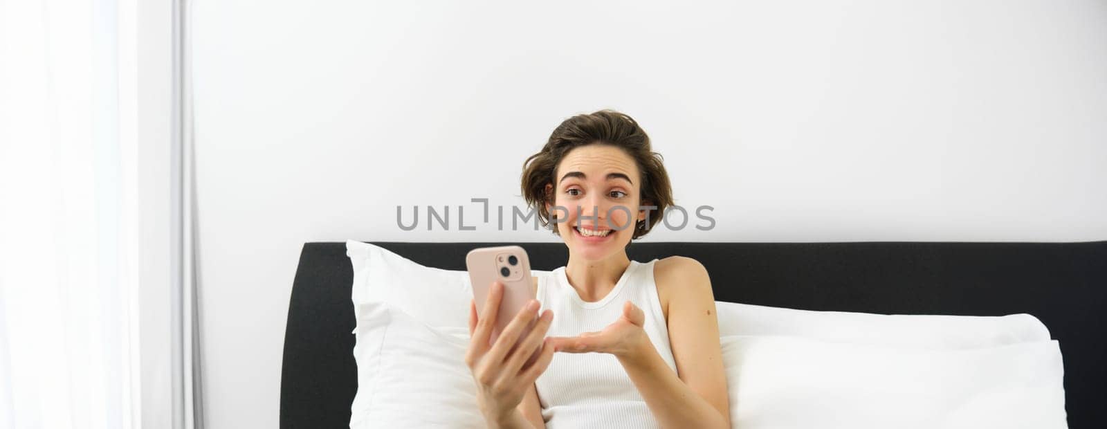 Cheerful smiling woman resting in bed, connects to online video call while lying in bedroom, talking to friend via smartphone app.