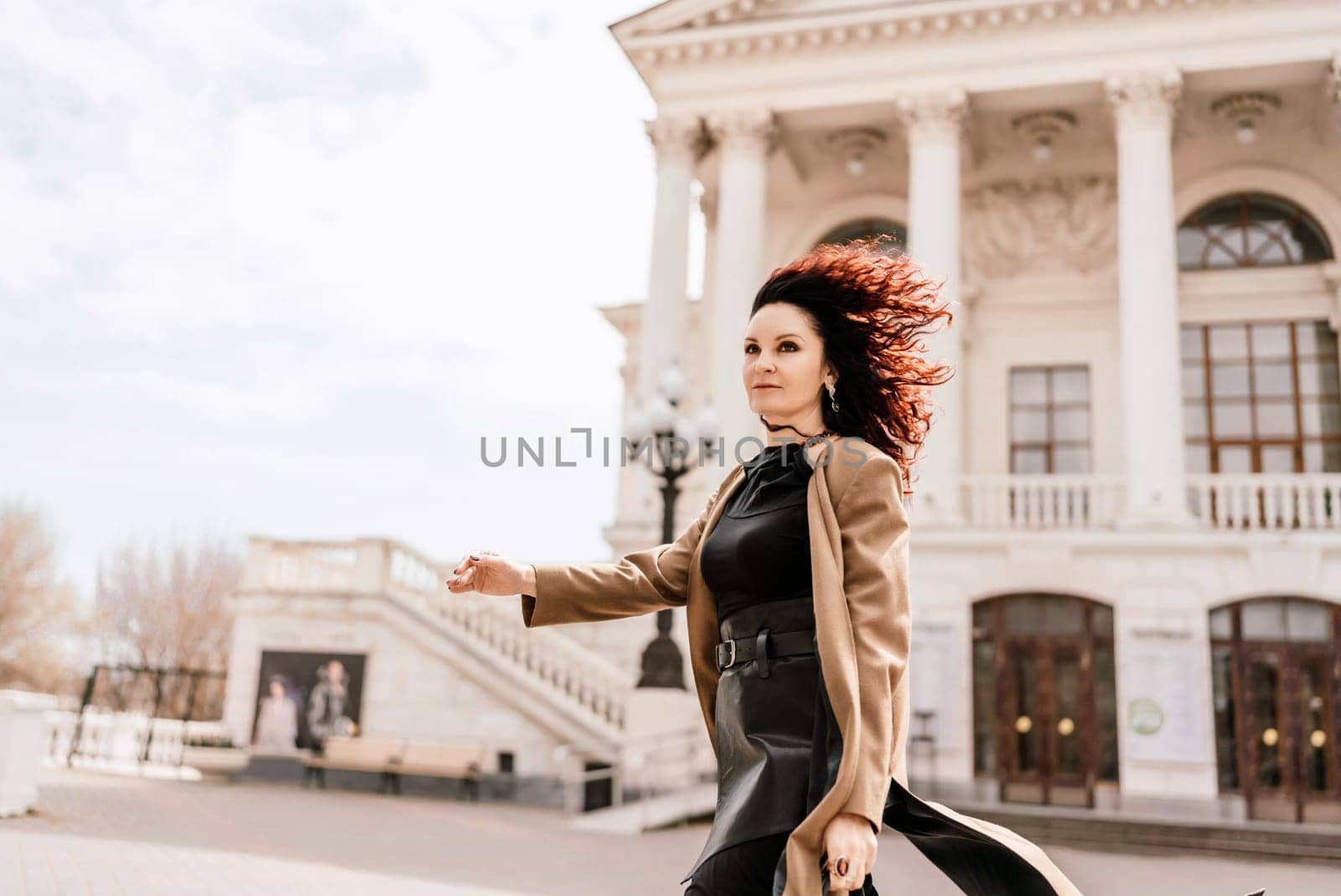 Woman street lifestyle. Image of stylish woman walking through European city on sunny day. Pretty woman with dark flowing hair, dressed in a beige raincoat and black, walks along the building