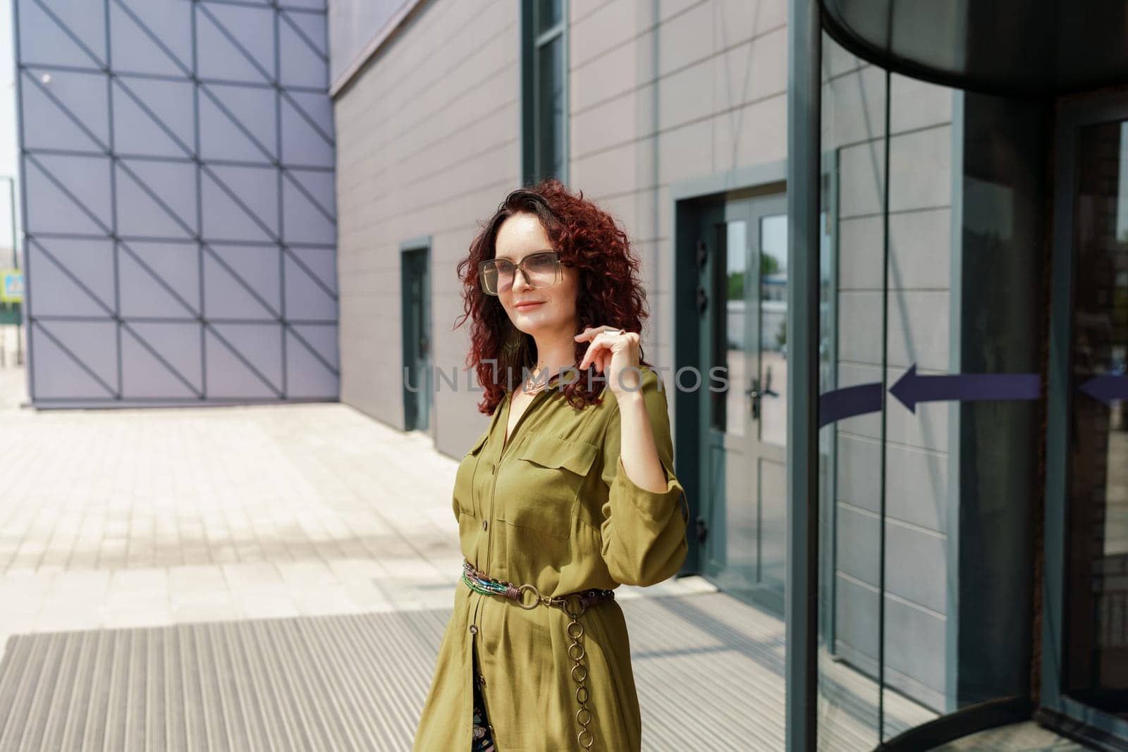 Portrait of a woman standing near a supermarket building. Caucasian model with long brunette hair, wears sunglasses and a khaki dress