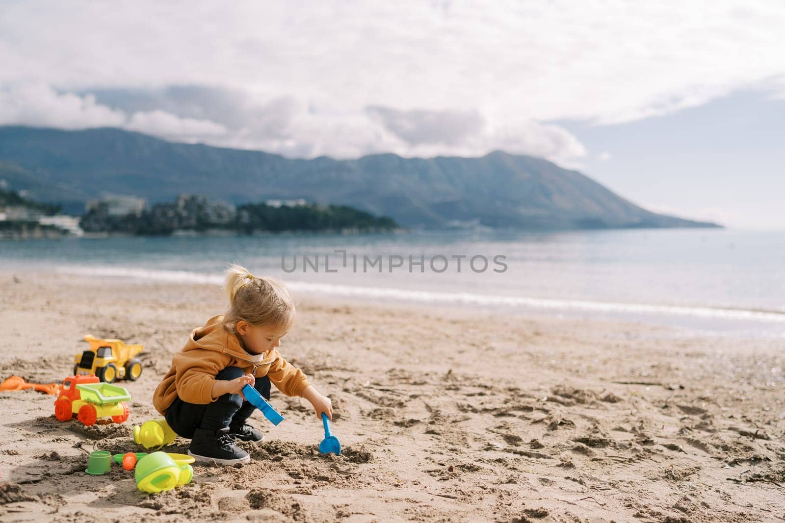 Little girl squats on the seashore and digs the sand with a shovel holding a plastic mold in her hand. High quality photo