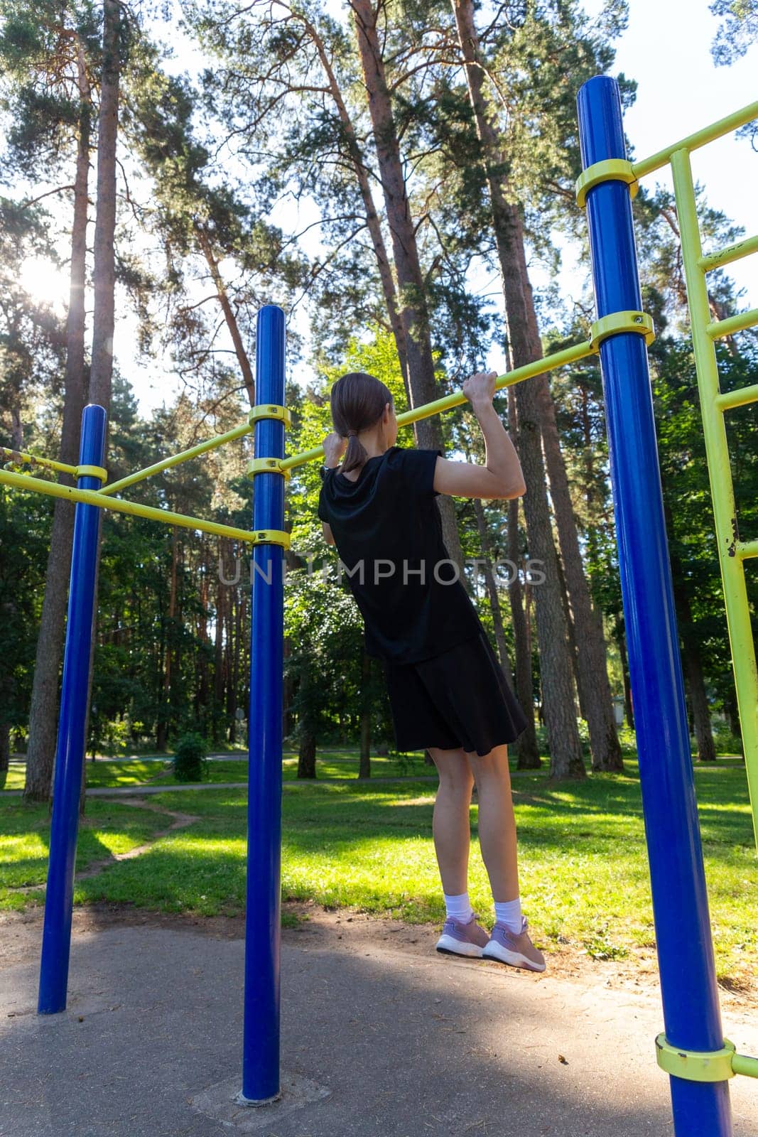 Portrait of beautiful teen age athletic girl training outdoors at open air gym at summer sunny day, gain hands muscles on horizontal bar. fitness, healthy lifestyle.
