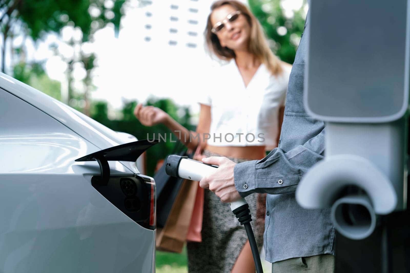 Young couple travel with EV electric car charging in green sustainable city outdoor garden in summer shows urban sustainability lifestyle by green clean rechargeable energy of electric vehicle innards