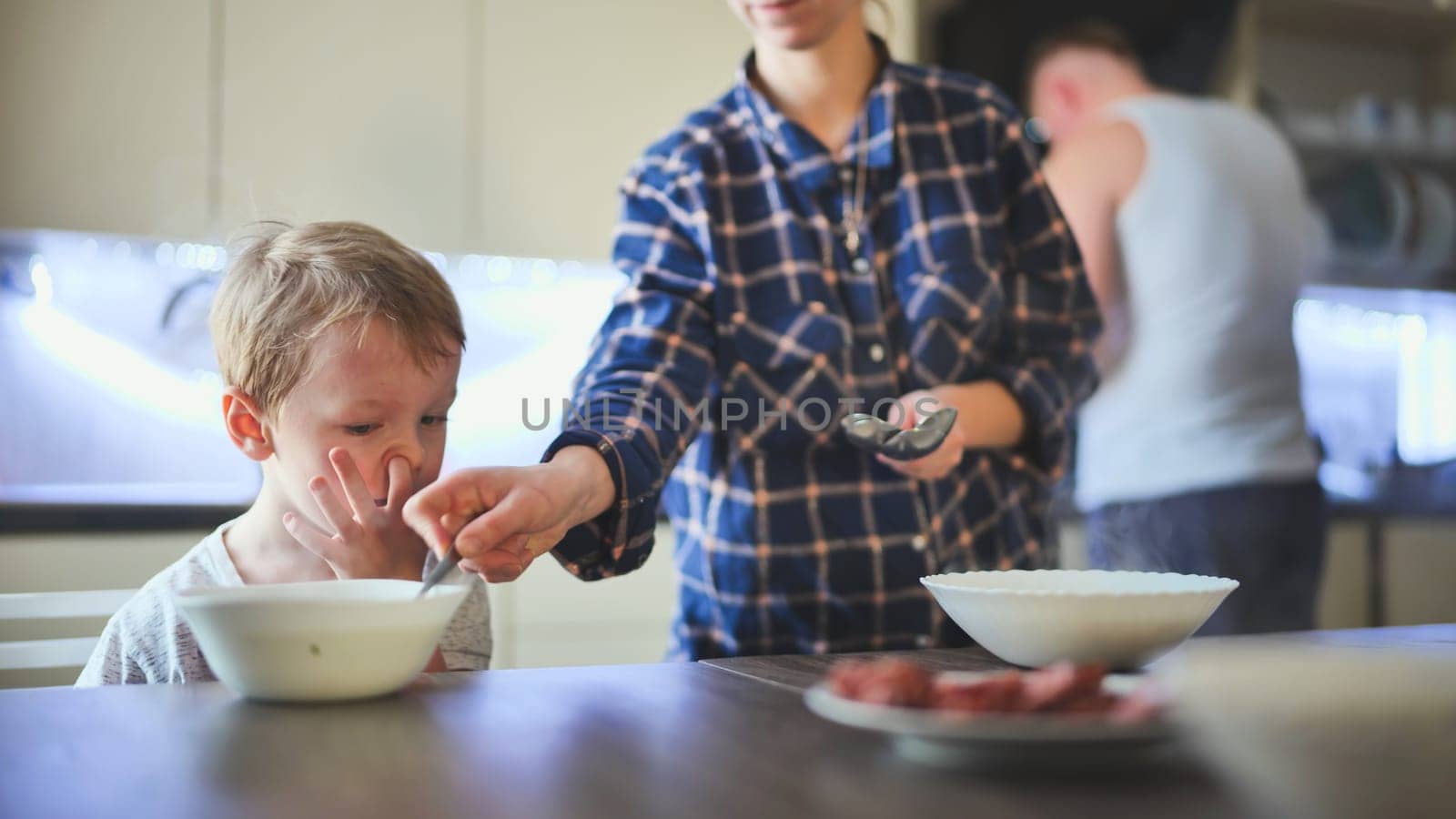 The child sits at the table in front of the bowl. The parents bring him a dish