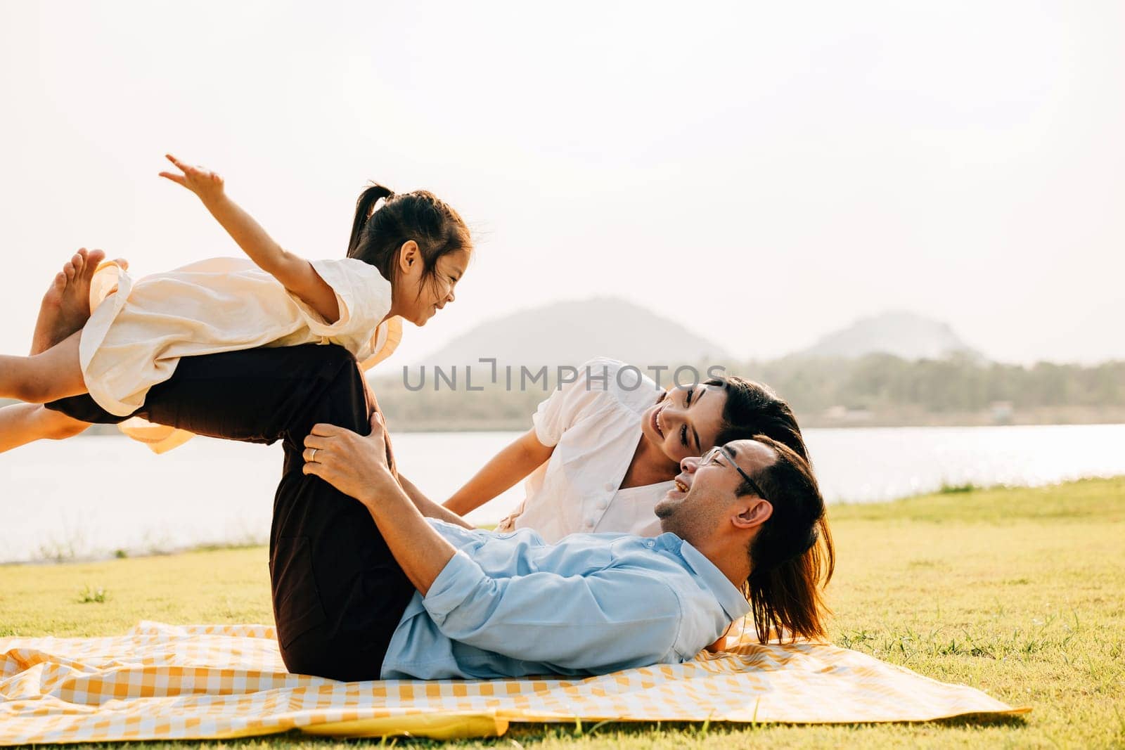 A playful family day in the park. Father lifts his daughter high, as she joyfully opens her arms and flies like a plane, while mother looks on with happiness. Family fun captured in a photograph