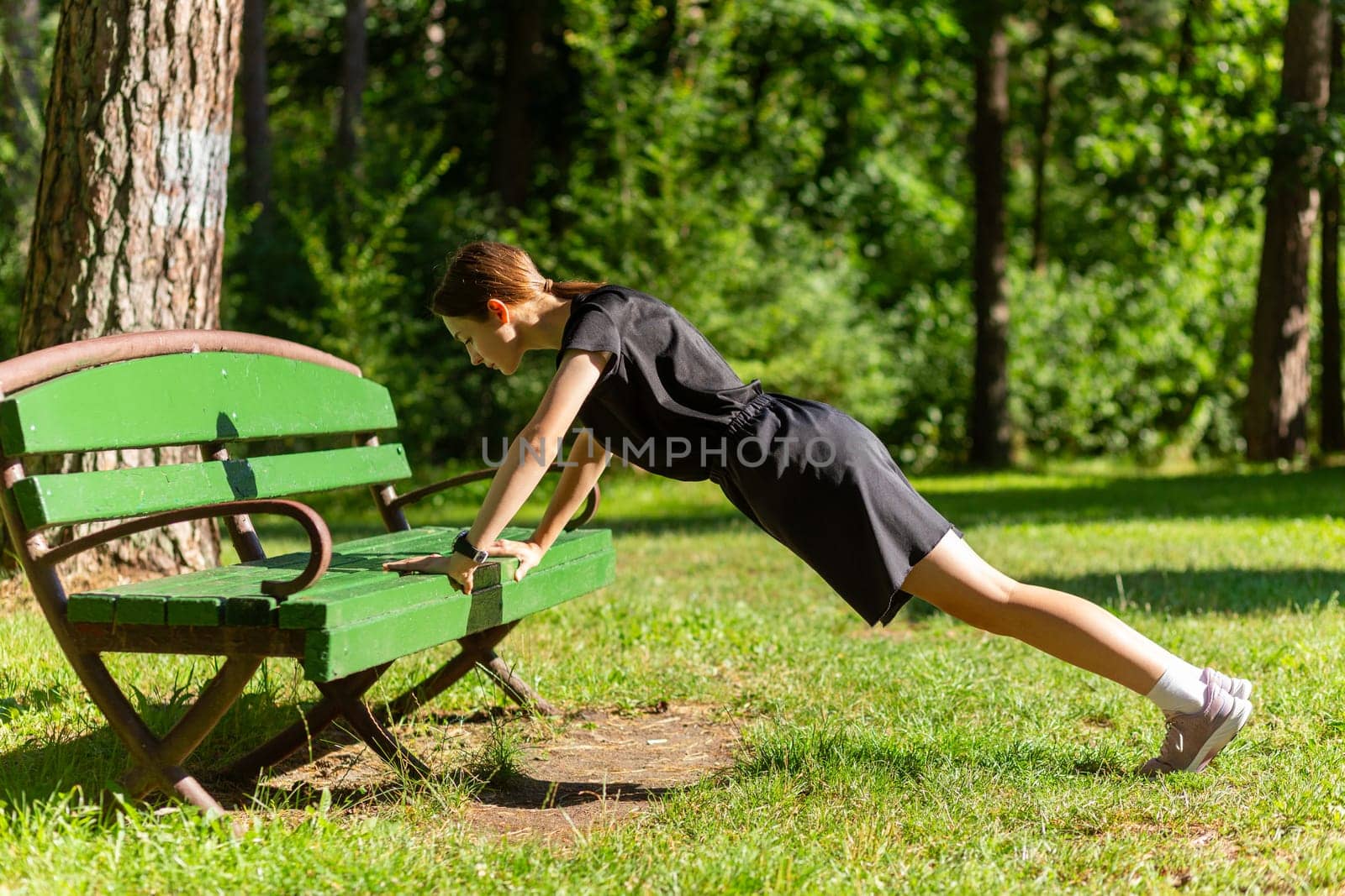 Beautiful young sporty woman in black t-shirt, black shorts and pink trainers warming up exercising triceps and chest dip doing push-ups from bench among trees before running.