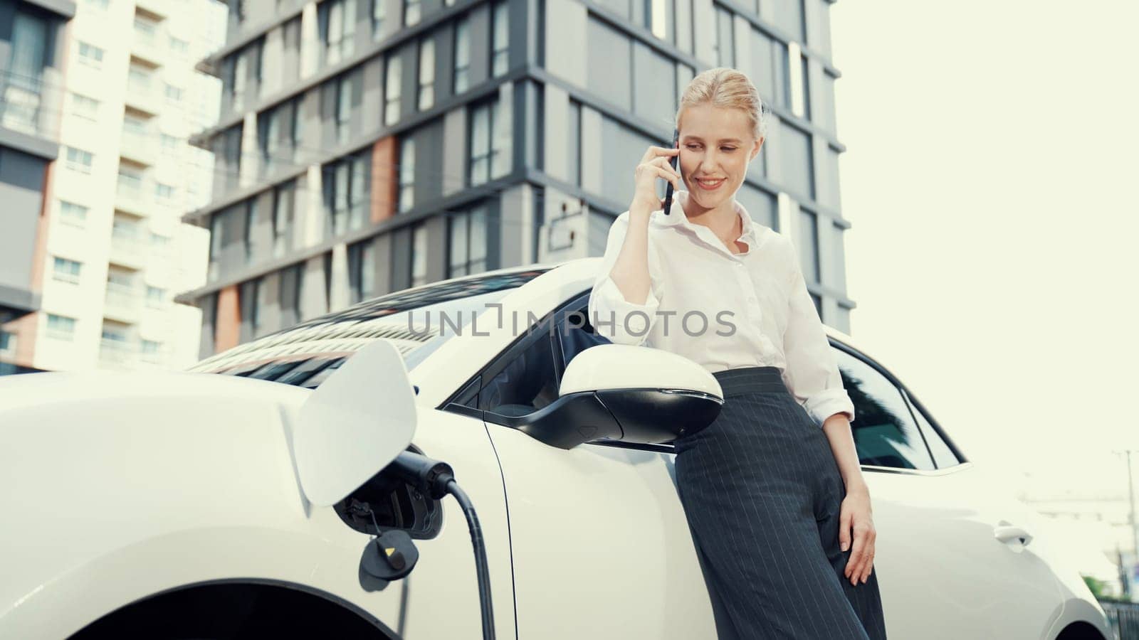 Progressive businesswoman leaning on electric car and charging station before driving around city center. Eco friendly rechargeable EV car powered by sustainable and clean energy.