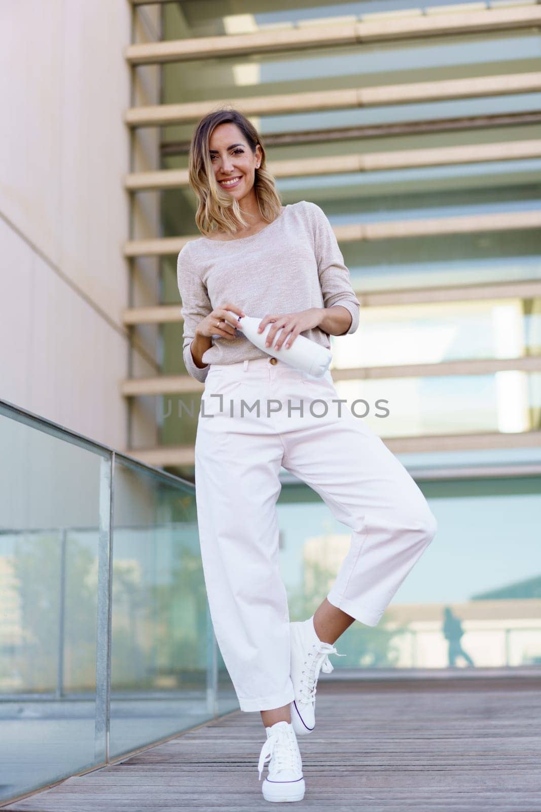 Full body happy female in stylish clothes, looking at camera with smile and opening bottle of water while balancing on one leg on path outside modern building