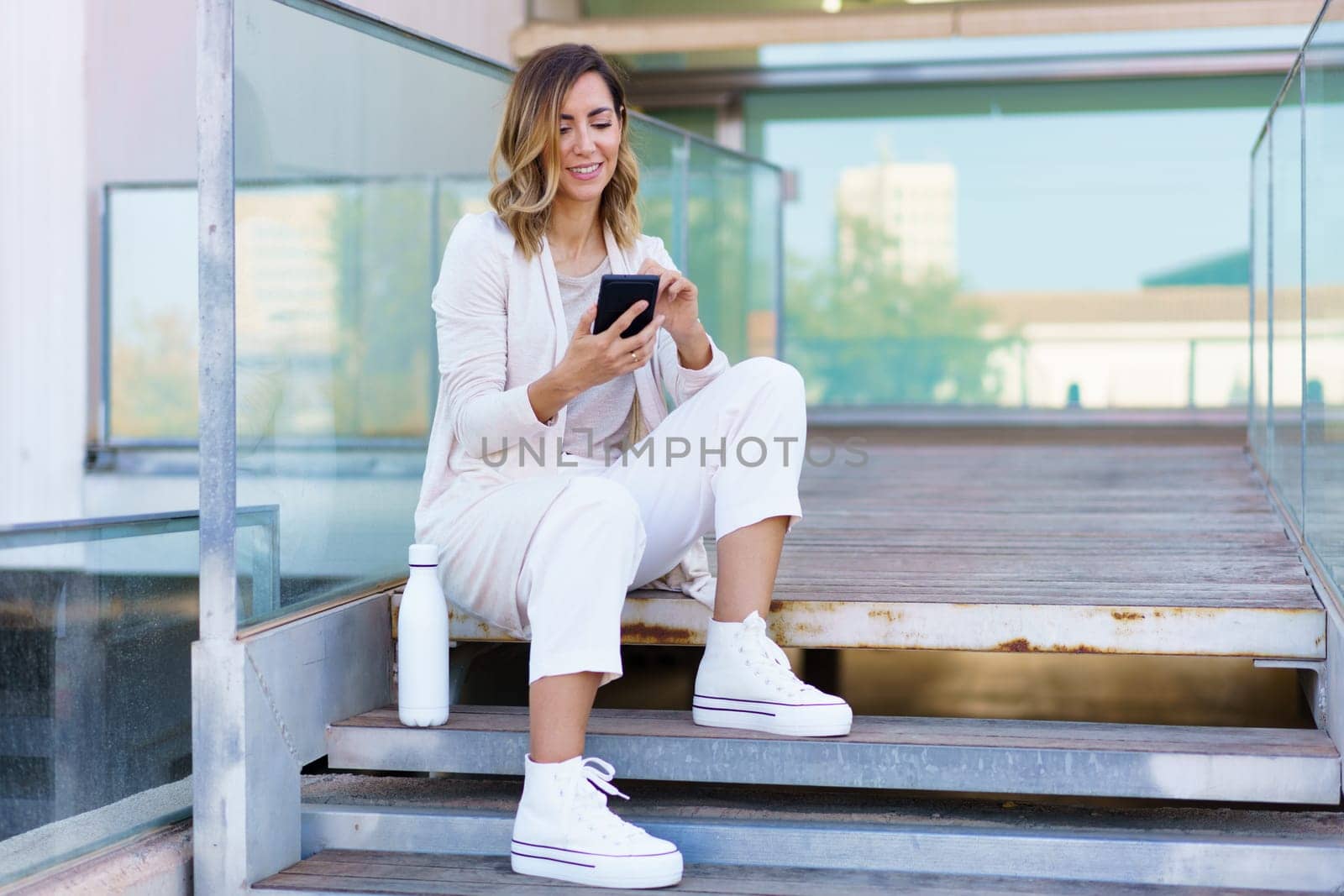 Middle aged woman taken a coffee break, sitting on steps near her office building with an eco-friendly ecological metal water bottle. Businesswoman using smartphone.